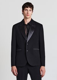 Schwarze Smokingjacke mit Satinrevers, zwei Knöpfen und kontrastierenden weißen Nähten an den Taschen, kombiniert mit einem schwarzen, transparenten Hemd darunter.