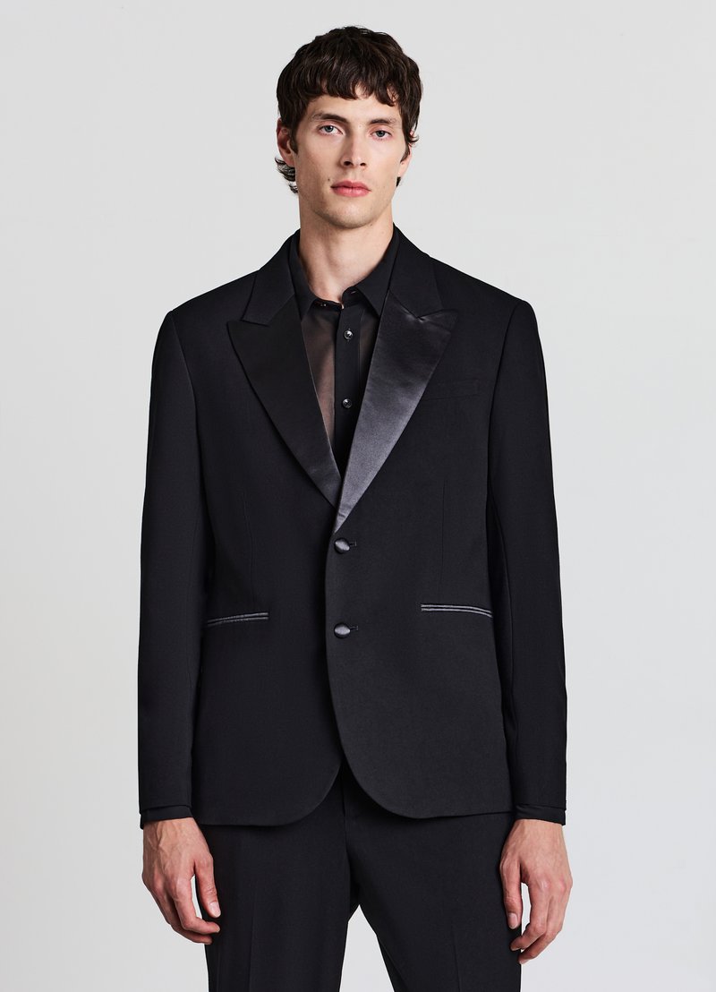 Schwarze Smokingjacke mit Satinrevers, zwei Knöpfen und kontrastierenden weißen Nähten an den Taschen, kombiniert mit einem schwarzen, transparenten Hemd darunter.