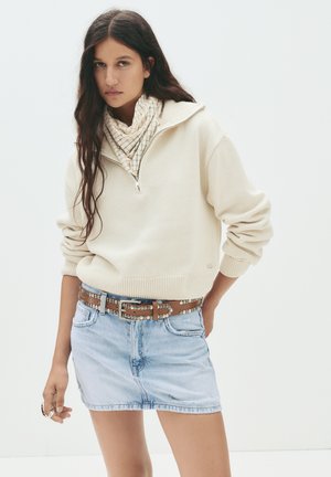 Pull en beige zippé avec des poignets côtelés, associé à une mini-jupe en denim bleu clair. Ceinture marron à clous et écharpe à motif à carreaux en accent.