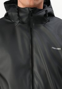 Giacca nera impermeabile con una texture liscia, dotata di una zip frontale e cappuccio regolabile. Logo del marchio "TRETORN" visibile in bianco.