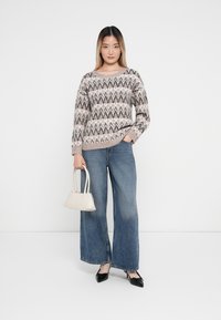Jasno beżowy sweter dzianinowy w zygzakowaty wzór, zestawiony z szerokimi dżinsami w niebieskim kolorze. Trzyma małą białą torebkę, czarne czółenka na szpilkach.