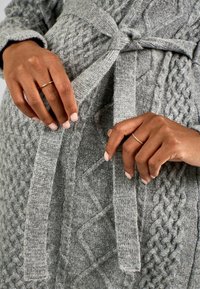Cardigan grigio a maglia con motivo a trecce e una cintura. Mani tengono il tessuto, mettendo in mostra la trama e i dettagli del design.