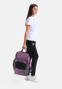 Zaino viola e nero con tasca frontale in rete, ampio scomparto principale e maniglia centrale per il trasporto, tenuto da una persona in abbigliamento sportivo.