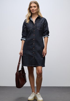 Mörk denimskjortklänning med knappstängning, uppvikt ärmar, två bröstfickor och en avslappnad passform. Matchas med en brun väska och vita sneakers.