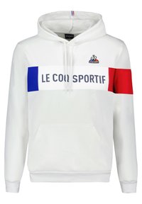 Un hoodie blanc avec une poche kangourou sur le devant, arborant une bande horizontale bleue et rouge avec le texte "LE COQ SPORTIF" et un logo brodé.