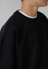 Persona che indossa un pullover nero a girocollo sopra una maglietta bianca, mostrata dalla spalla in su con uno sfondo grigio neutro.