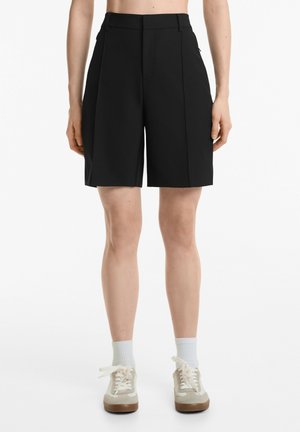 Shorts noirs ajustés à taille haute, avec un devant plissé, des poches latérales et une coupe droite, fabriqués en tissu lisse.