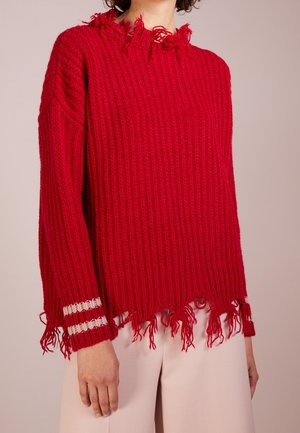 Pull en tricot rouge avec bords usés à l'ourlet, aux poignets et au col, avec deux bandes blanches sur le poignet gauche, porté avec un pantalon clair.
