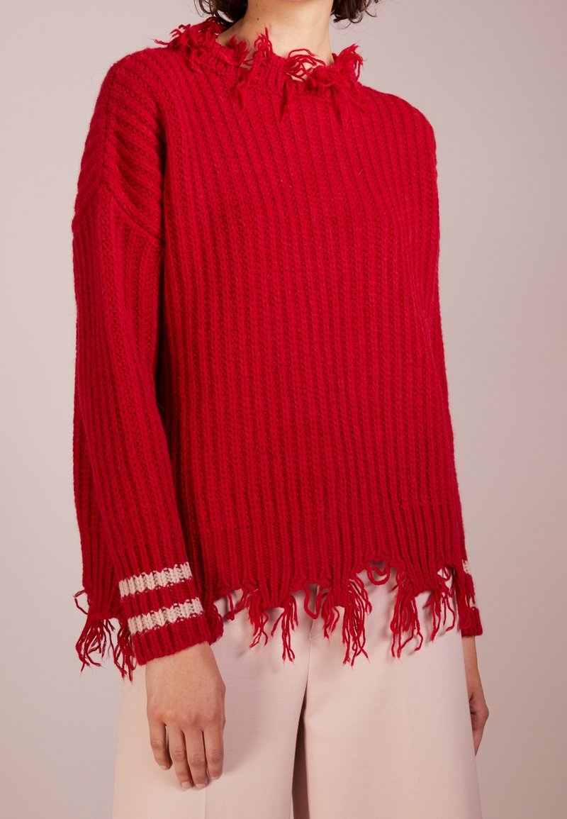 Pull en tricot rouge avec bords usés à l'ourlet, aux poignets et au col, avec deux bandes blanches sur le poignet gauche, porté avec un pantalon clair.