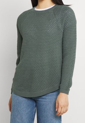 Jersey verde de punto con un patrón texturizado, puños y dobladillo acanalados. Presenta un cuello redondo y un ajuste holgado, combinado con vaqueros azules.