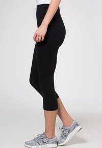 Černé vysoké capri kalhoty vyrobené z elastického materiálu, s hladkou texturou a přiléhavým střihem. V kombinaci se šedými sportovními botami.