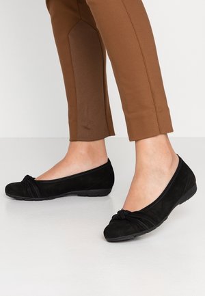 Czarne zamszowe baletki z detalem w formie skręconego węzła na palcu i niską, elastyczną gumową podeszwą, noszone z brązowymi spodniami sięgającymi kostek.