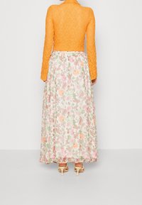 Top corto de encaje naranja con mangas largas, combinado con una falda larga de patrón floral en tonos pastel y sandalias de tacón beige.