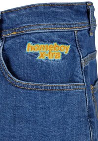Denim farkut keski sinisessä, joissa on oranssit tikit ja brodeerattu logo, joka lukee "homeboy x-tra" takataskussa.