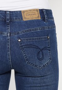 Jean en denim avec un lavage bleu foncé, mettant en avant une écusson logo sur la taille et des coutures détaillées sur les poches arrière.