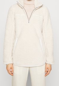 Valkoinen sherpa-fleecepullover, jossa neljänneksen vetoketju ja sivutaskut, ylle puettu beigen värisen pooloneuleen kanssa, yhdistettynä valkoisiin housuihin.