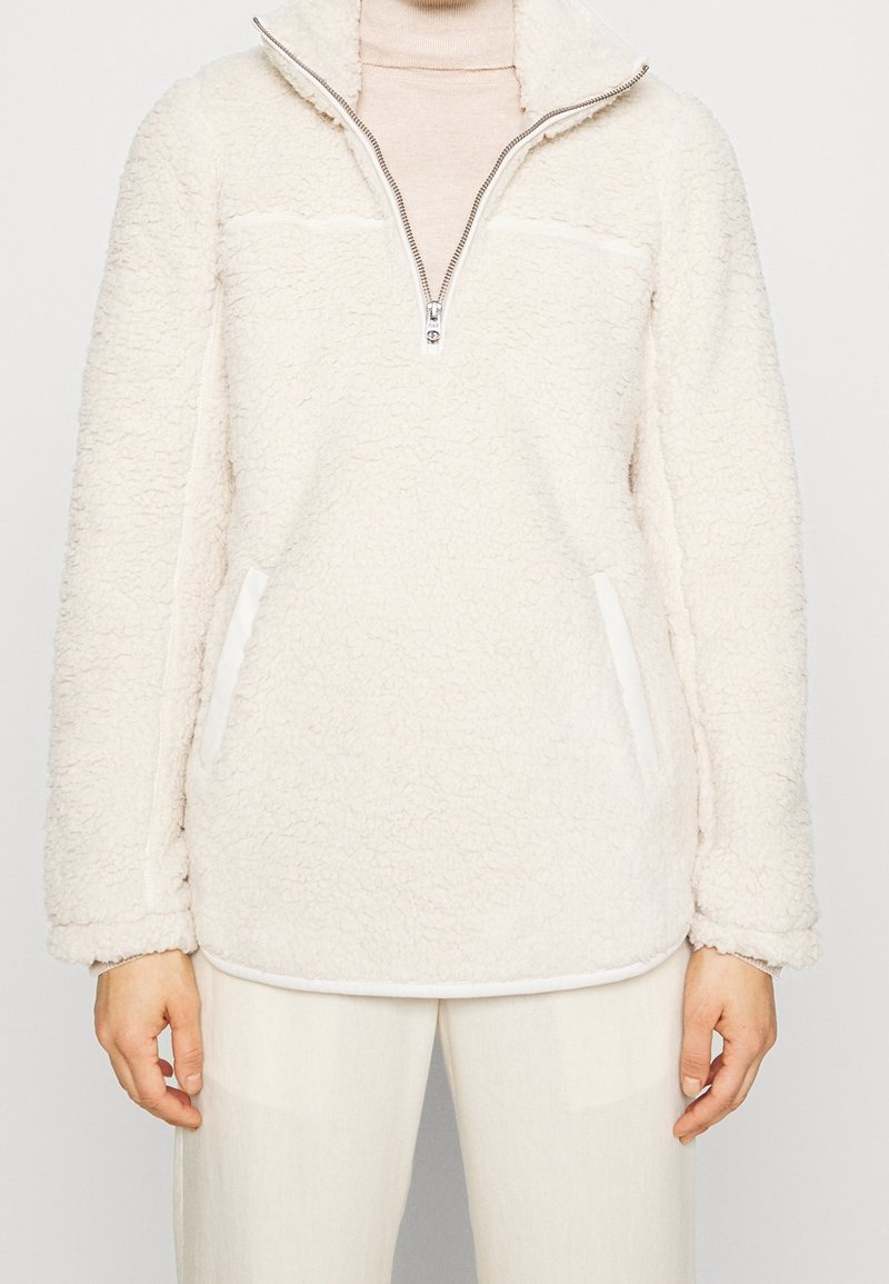 Valkoinen sherpa-fleecepullover, jossa neljänneksen vetoketju ja sivutaskut, ylle puettu beigen värisen pooloneuleen kanssa, yhdistettynä valkoisiin housuihin.