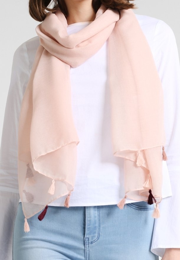 Écharpe rose clair avec des pampilles drapée autour du cou d’une personne, associée à une chemise blanche à manches longues et un jean bleu clair.