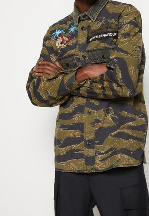 Homme, bras croisés, portant une chemise camouflage verte et noire avec une broderie de palmier et de crâne ainsi qu'un écusson texte « Brave Behaviour ».