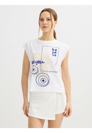 Joven con cabello castaño recogido en una coleta lleva una camiseta sin mangas blanca con gráfico y una minifalda blanca asimétrica sobre un fondo liso.