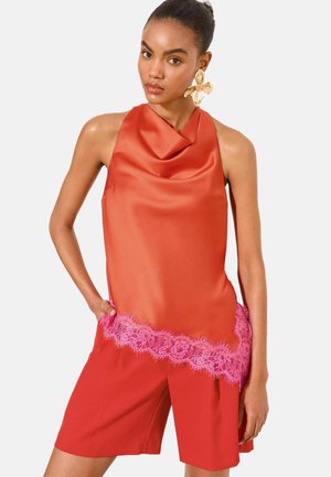 Woman wearing sleeveless orange satin top with pink lace trim and matching red shorts, accessorized with large gold flower earring.