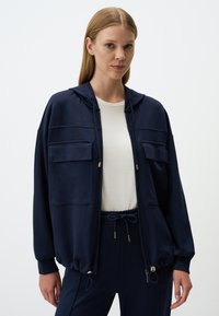 Gilet zippé bleu marine avec deux poches poitrine, capuche réglable avec cordons de serrage et poignets élastiques. Tissu doux avec une texture lisse.