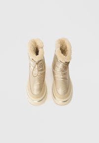 Gold puffer-style boots with a fluffy cream fur lining, round toe, and a textured rubber sole. Features a pull tab for tightening.