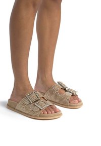 Beige schuifsandalen met versierde studs, twee grote gespaccenten en een suède textuur, met een open teen ontwerp.