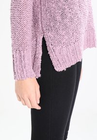 Pletený svetr v levandulové barvě s žebrovanými detaily, vybavený postranním rozparkem a volným střihem. V kombinaci s černými legínami. Viditelný texturovaný detail látky.