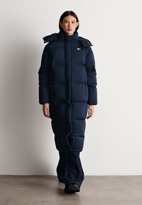 Námornícky puffový kabát s prešívaným dizajnom, vysokým golierom a nastaviteľnou kapucňou, kombinovaný s tmavými nohavicami a čiernymi topánkami na neutrálnom pozadí.
