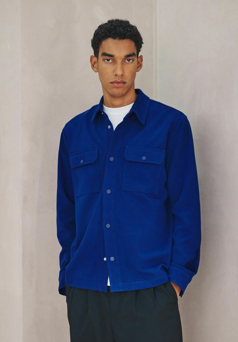 Blaues Hemd mit Knöpfen und zwei Brusttaschen, aus glattem Stoff, mit spitzem Kragen und lässiger Passform, kombiniert mit schwarzen Hosen.