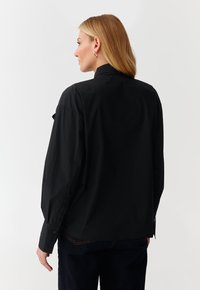 Camicia nera con colletto realizzata in tessuto liscio; presenta maniche lunghe e polsini abbottonati. La vista posteriore mostra una vestibilità comoda e un design minimale.
