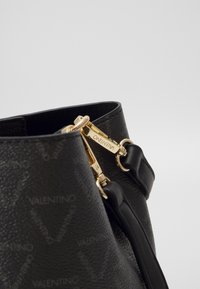 Närbild på svart Valentino-läderhandväska med guldfärgade dragkedjehandtag ingraverade med Valentino-logotyp och tonalt logotypmönster på lädret.