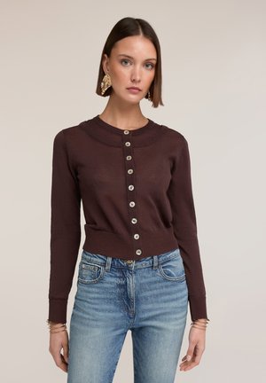 Nainen, jolla on päällään tummanruskea napitettu neuletakki, siniset farkut, kultaiset lehtimuotoiset korvakorut ja useita kultaisia rannekoruja beigen taustan edessä.