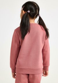 Felpa rosa realizzata in un morbido tessuto testurizzato. Polsini e orlo a costine, vestibilità leggermente ampia, indossata sopra pantaloni abbinati. Nessun logo o motivo visibile.