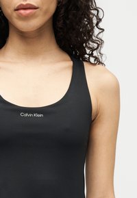 Κοντινό πλάνο ενός ατόμου που φοράει μαύρο αμάνικο τοπ Calvin Klein, με εμφανείς λεπτομέρειες από τον λαιμό, τον ώμο και το πάνω μέρος του στήθους.