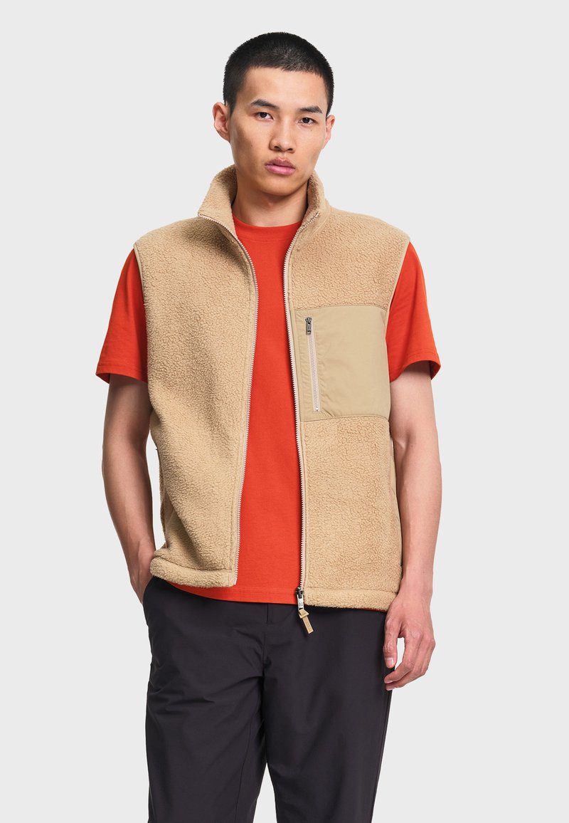 Homme portant un gilet polaire beige zippé sur un t-shirt orange à manches courtes et un pantalon noir devant un fond clair uni.