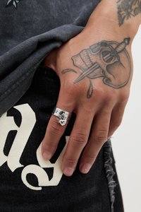 Sølvring med ilddesign på en finger, satt mot et svart plagg med hvit skrift og synlige tatoveringer på hånden.