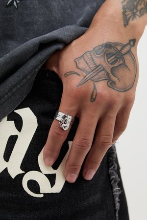 Bague en argent avec un motif de flamme sur un doigt, placée sur un vêtement noir comportant des lettres blanches et des tatouages visibles sur la main.