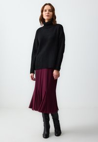Pull à col roulé noir surdimensionné, associé à une jupe midi plissée prune. Le mannequin porte des bottes noires jusqu'aux genoux. Fond clair.