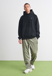 Ung mann står iført svart Nike hettegenser, olivengrønne joggebukser og hvite sneakers på grønt gulv mot hvit vegg.