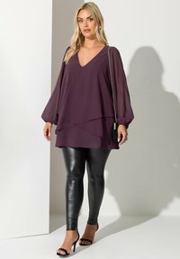 Blusa viola a strati con scollo a V, maniche ampie; abbinata a leggings in pelle nera aderenti e sandali neri con punta aperta.