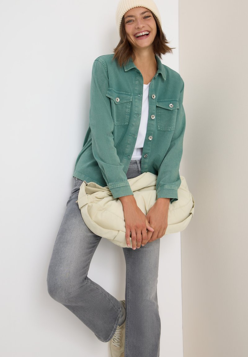 Femme souriante portant un bonnet en tricot beige, une veste verte à boutons, un jean gris et des baskets beiges, tenant un grand sac rembourré blanc cassé.