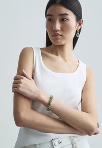Groene edelsteenarmband met gouden accenten, voorzien van ingewikkelde schakels en een glanzende afwerking, gedragen door een model met donker haar.