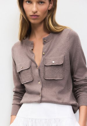 Frau trägt mauvefarbene, geknöpfte Strickjacke mit großen Fronttaschen, kombiniert mit einem weißen Rock, vor einem schlichten hellen Hintergrund.