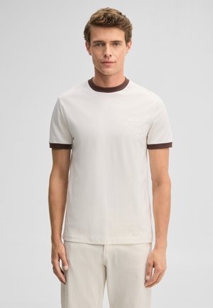 Mann steht, trägt ein weißes T-Shirt mit dunkelbraunem Kragen und Ärmelbündchen, schaut nach vorne vor einem schlichten hellen Hintergrund.