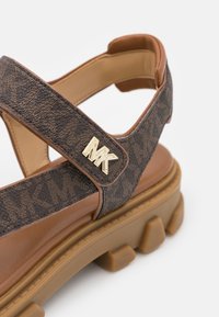 Sandale marron à semelle épaisse, logo doré de la marque "MK" sur une bride réglable à motif, et semelle intérieure rembourrée en gros plan.