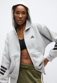 Γκρι φούτερ με φερμουάρ, κατασκευασμένο από μαλακό ύφασμα, με ρυθμιζόμενη κουκούλα, μπροστινές τσέπες και μαύρες τρεις ρίγες στα μανίκια.