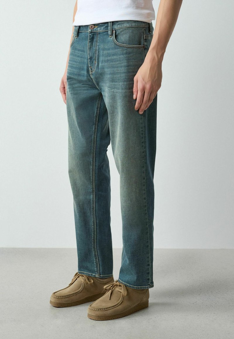 Hombre vestido con vaqueros azules desgastados y zapatos beige de gamuza con cordones, de pie frente a un fondo liso de color claro.