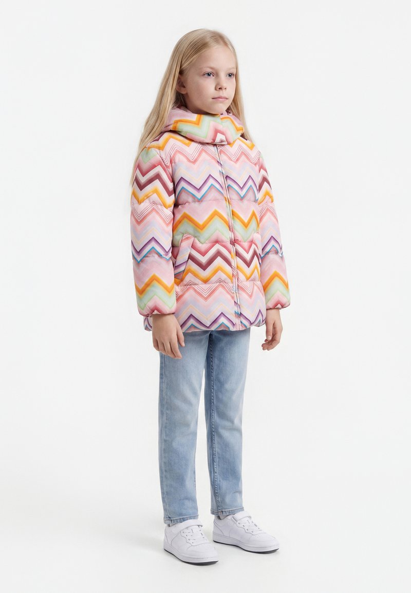 Jeune fille debout portant une doudoune colorée à motif zigzag, un jean bleu clair et des baskets blanches sur fond blanc.